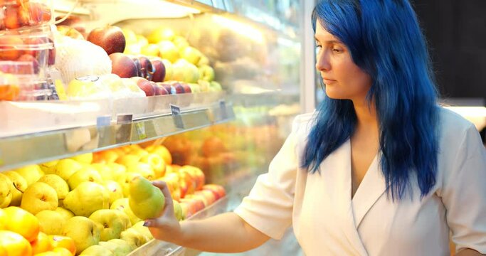 Young woman choosing imported pears in big Asian grocery store. She take one fruit from heap on shelf, turn it in hand and look around, put back if not satisfied, or place in bag to buy.