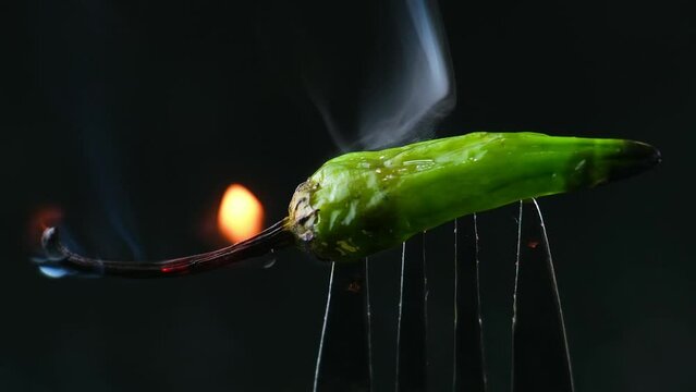 Burning spicy Hot Green chili pepper on a fork in flames on a black background. Spicy food concept.
