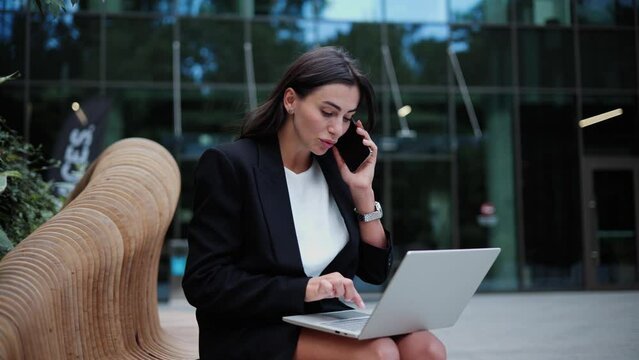 Young beautiful business girl uses laptop sitting on bench outside while talking on mobile phone. Busy young woman with laptop having online conversation chatting with coworkers sending message.