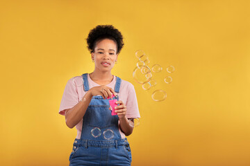 Woman, blowing bubbles and fun portrait in studio while playful for birthday, space or party celebration. Black female model person with liquid soap, natural beauty and fashion on a yellow background - Powered by Adobe