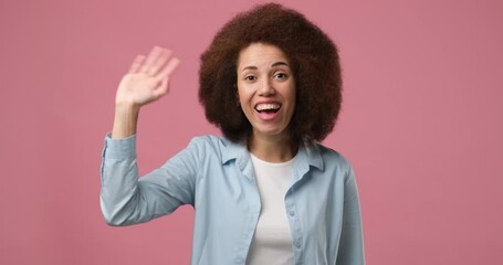 Happy friendly young african american woman waves palm in hello gesture welcomes someone with hospitable expression expresses positive emotions standing over pink background
