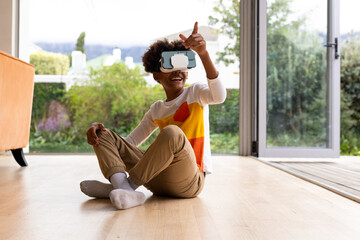 Happy african american boy sitting on floor using vr headset and touching virtual screen