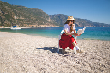 happy girl in a red skirt on an empty beach sits on a chair and reads a book