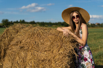 beautiful glamorous girl in a dress and a straw hat on a field near a haystack.