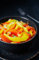 Salad with red and yellow roasted pepper slices, garlic and parsley in a black ceramic bowl on a black background. Concept of healthy eating. Traditional Italian dish. Vegetarian and vegan food.