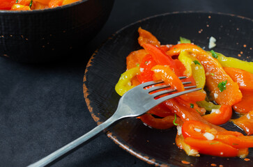 Salad with red and yellow roasted pepper slices, garlic and parsley in a black ceramic plate on a black background. Concept of healthy eating. Traditional Italian dish. Vegetarian and vegan food.