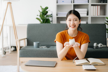 Asian women's hand saving a coin into piggy bank with wooden to calculateon and tablet on the table for business, finance, saving money and property investment concept.