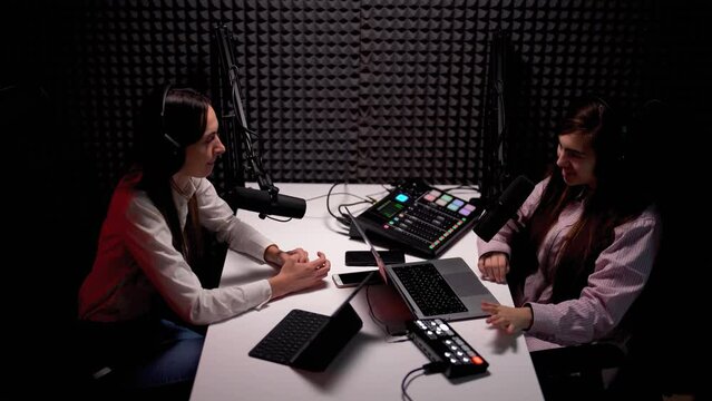 View from above of young woman podcaster interviewing her female guest, both wearing headphones and speaking into microphones in a modern acoustic studio 