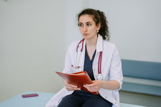 Puzzled female doctor with phonendoscope on neck holds folder with histories of sickness  looks away with pensive face expression. Tired nurse sitting on medical bench at exam room.