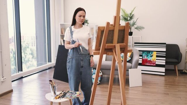 Full length view of barefoot woman in denim overalls working on easel in spacious art studio on sunny day. Confident adult focusing on paint application using wide brush in comfortable interior.