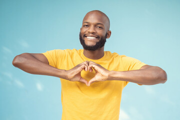 Hand, heart and portrait of man in studio for love, happy and smile against a blue background...