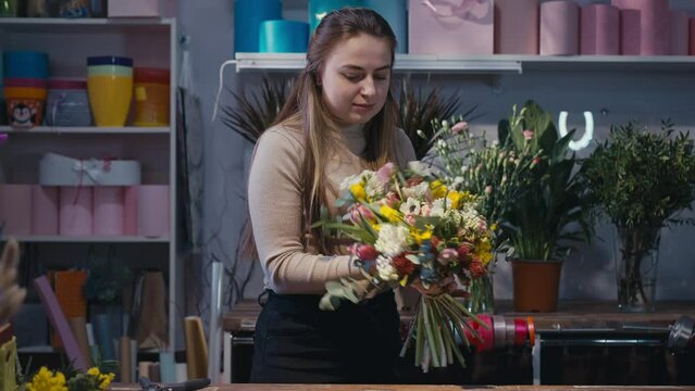 Happy confident young woman cutting flower stems bottom stretching bouquet to camera smiling. Medium shot portrait of positive Caucasian florist bragging posing in flower shop indoors