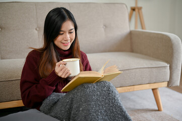 A beautiful Asian woman in a cozy sweater is sipping hot chocolate and reading a book