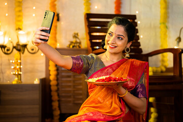 Happy indian girl in saree taking selfie on mobile phone by holding flowers plate during festival...