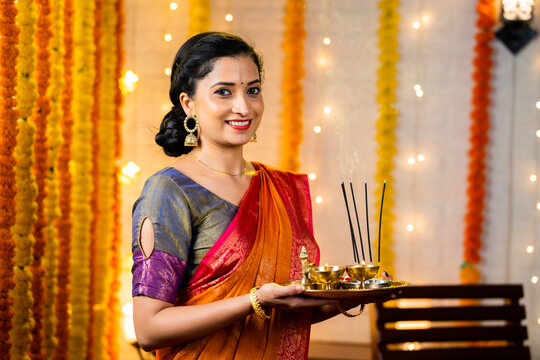 Happy indian girl in traditional ethnic wear holding pooja plate with incense sticks looking camera during diwali festival celebration at home - concept Indian culture, positive emotion and happiness