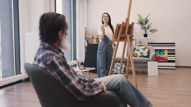 Attractive brunette lady in denim overalls sketching on canvas while looking at older man sitting in chair at workshop. Young caucasian artist depicting live model full body in art studio interior.
