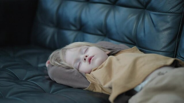 Witness the restful nap of a 3-year-old Caucasian boy in a medium shot, as he sleeps serenely on a black leather couch. A charming moment of peaceful relaxation.