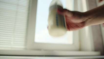 Stirring and shaking a bottle of milk baby food against the background of a window. Preparing infant formula. Copy space for text