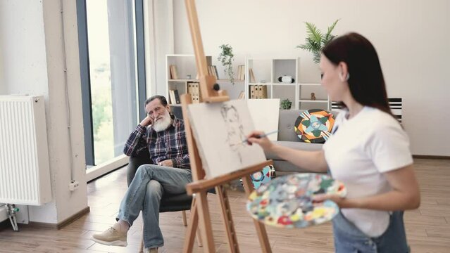 Relaxed older man posing in comfortable chair while young woman with palette applying paint on canvas. Avid arts student making initial drawing during portrait painting lesson in studio space.