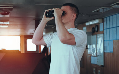 Deck officer on the bridge during navigational watch at sea. Offshore technician. Seafarer. Seaman. Navigator. A man in the wheelhouse during watchkeeping duty. Look out with binoculars. © I am from Mykolayiv