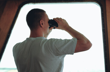 Deck officer on the bridge during navigational watch at sea. Offshore technician. Seafarer. Seaman. Navigator. A man in the wheelhouse during watchkeeping duty. Look out with binoculars. © I am from Mykolayiv