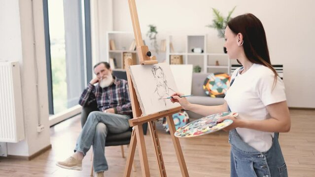 Self-employed female painter in denim overalls depicting sitter head-and-shoulders in modern studio space. Attractive brunette lady sketching out rough ideas of senior man's portrait on canvas.