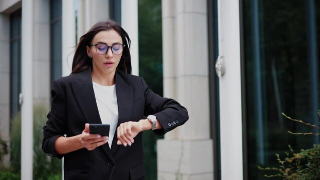 Young businesswoman is in a rush to a meeting, checking her watch near a modern office building. Busy female office worker, dressed in a classic suit and glasses, is running late for work outdoors.