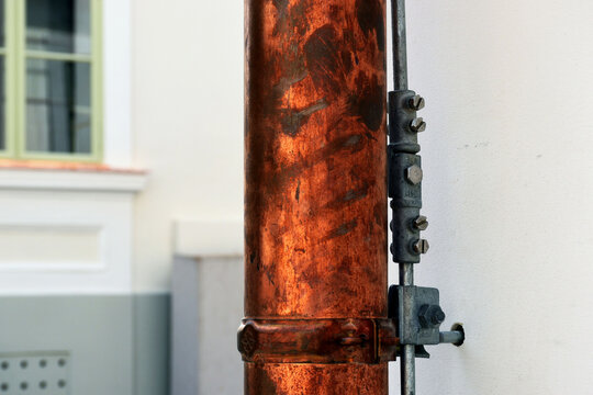 shiny new copper rain water leader, downspout or down pipe detail. lightning protection grounding rod. home renovation, construction concept. textured white stucco exterior wall and window background