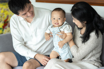 Young and cheerful mother and father sitting on couch relaxing while playing with cute little new born baby boy and toddler who is trying to stand with support at home during weekend