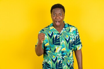 Young latin man wearing hawaiian shirt over yellow background angry and mad raising fist frustrated...