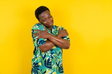 Young latin man wearing hawaiian shirt over yellow background Hugging oneself happy and positive,...