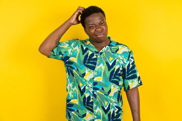 Young latin man wearing hawaiian shirt over yellow background being confused and wonders about something. Holding hand on head, uncertain with doubt. Pensive concept. - obrazy, fototapety, plakaty