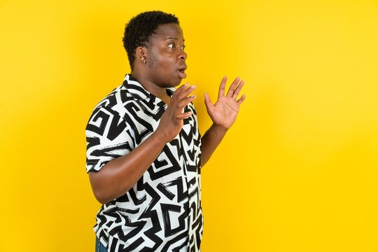 Young latin man wearing printed shirt over yellow background shouts loud, keeps eyes opened and hands tense.