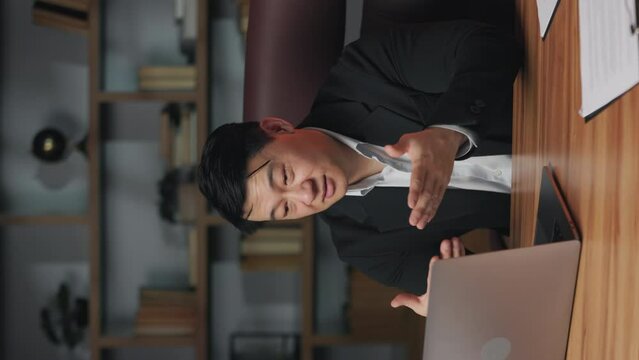 Experienced asian worker in formal outfit having online conversation using laptop webcam in office. Confident employer conducting job interview with candidate via internet connection. Vertical video.