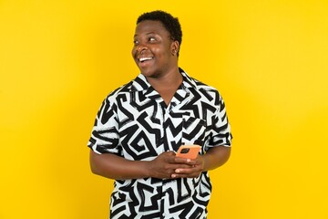 Young latin man wearing printed shirt over yellow background holding a smartphone and looking sideways at blank copyspace.