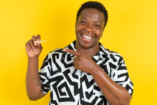  Young latin man wearing printed shirt over yellow background holding an invisible aligner and pointing at it. Dental healthcare and confidence concept.