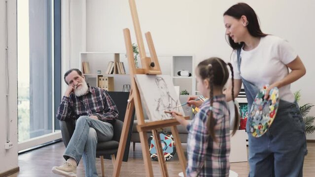 Friendly elderly man in jeans waving hello to little girl standing near female artist with palette in home studio. Smiling pensioner in soft chair greeting granddaughter on art lesson with mother.