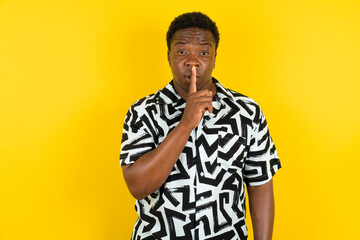 Surprised  Young latin man wearing printed shirt over yellow background makes silence gesture, keeps finger over lips and looks mysterious at camera