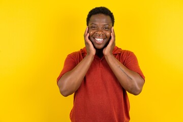 Happy Young latin man wearing red t-shirt over yellow background touches both cheeks gently, has tender smile, shows white teeth, gazes positively straightly at camera,