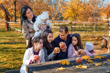 big family has picnic in autumn city park, children and parents sitting together at the table, with apples and yellow maple leaves, happy people enjoying beautiful nature, bright sunny day