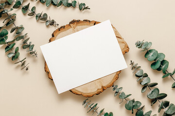 Aesthetic blank paper mockup card on wooden stand with eucalyptus leaves on beige background, top view. Paper card template for wedding, greeting, invitation and branding, bohemian style