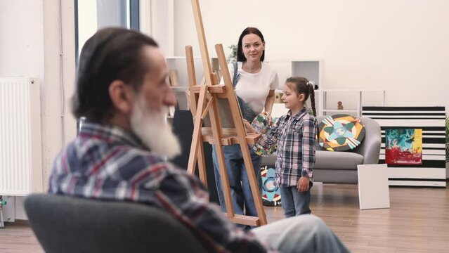 Selective focus of caucasian ladies in denim outfits painting on canvas while older male posing in chair on foreground. Inspired mother and daughter drawing image of calm grandad in art classroom.