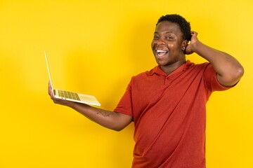 Photo of amazed Young latin man wearing red t-shirt over yellow background holding modern gadget...