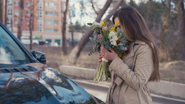 Smiling young woman finding bouquet on car hood on urban city street. Portrait of happy satisfied Caucasian lady with Valentine's surprise flowers outdoors