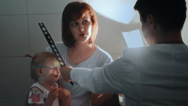 Diagnostic procedure skiascopy with pediatric ophthalmologist. Optometrist illuminates pupil of eye with mirror of ophthalmoscope, skiascopic ruler. Checking vision of little girl kid with eye disease