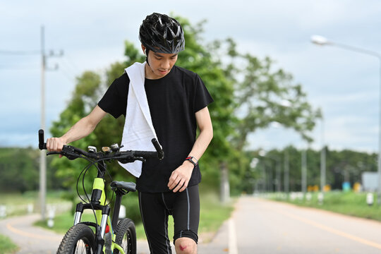 Young asian male cyclist with injured knee carry bicycle walk in park after an accident