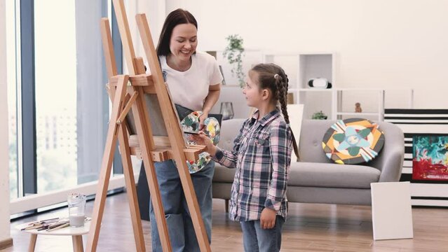 Portrait of beautiful female and little child in casual clothes standing behind easel with painting in workshop. Attentive art person teaching inquisitive small student mixing oil paints on paper.