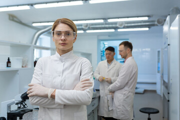 Portrait of serious mature woman scientist, group leader research boss standing inside laboratory wearing white medical coat and protective glasses and looking at camera.