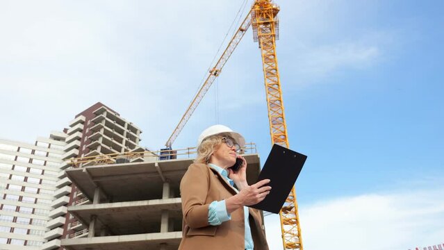 Businesswoman talks on smartphone with client standing against construction site and blue sky. Manufacturing industry and building construction low angle shot