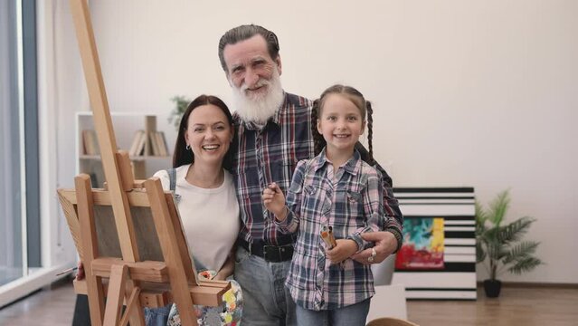 Portrait of proud older adult cuddling beautiful lady with palette and cute girl with brushes in home interior. Three generations of painters smiling happily at camera with artworks in background.